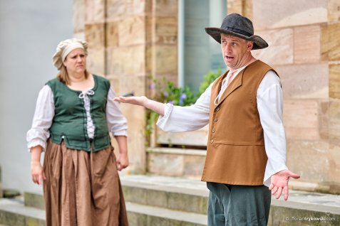 Zwei Schauspieler in historischer Kleidung stehen vor einem Gebäude, einer gestikuliert mit ausgestrecktem Arm, die andere steht im Hintergrund