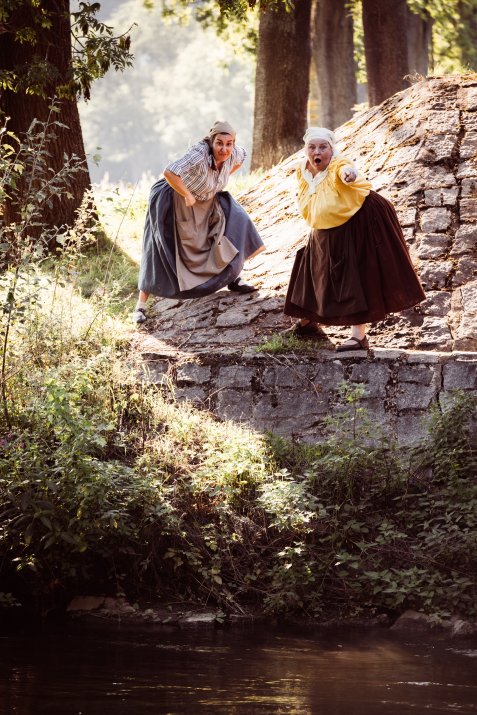 Zwei Fischweiber stehen am Flussufer auf einer steinernen Böschung und entdecken Fische.