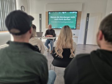 Menschen sitzen in einem Tagungsraum vor einem Vortragenden, im Hintergrund ein großer Bildschirm mit dem Text 'Warum die Nürnberger nicht nach Fürth durften & andere Kirchweihgeschichten'.
