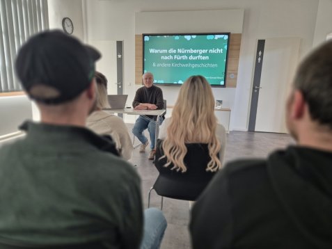 Menschen sitzen in einem Tagungsraum vor einem Vortragenden, im Hintergrund ein großer Bildschirm mit dem Text 'Warum die Nürnberger nicht nach Fürth durften & andere Kirchweihgeschichten'.