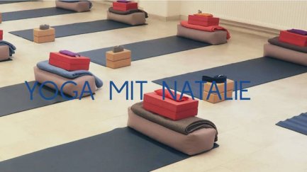 Mehrere Yogamatten mit gefalteten Decken und gestapelten Blöcken in einem hellen Raum, Schriftzug 'YOGA MIT NATALIE' sichtbar.