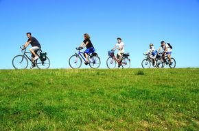 Fünf Personen fahren hintereinander auf Fahrrädern auf einem grasbewachsenen Hügel unter blauem Himmel