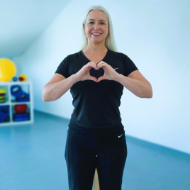 Frau in schwarzem Sportoutfit formt mit den Händen ein Herz vor dem Oberkörper in einem hellen Raum mit Sportgeräten im Hintergrund