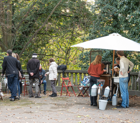 Besucher:innen auf dem Friedhofscafé mit Bäumen im Hintergrund, einige an einem Stand mit Sonnenschirm.