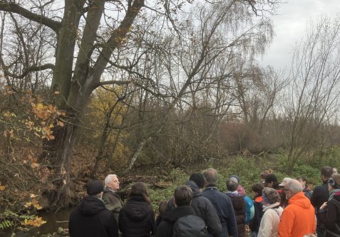 Gruppe von Menschen steht an einem Bachufer mit kahlen Bäumen im Hintergrund
