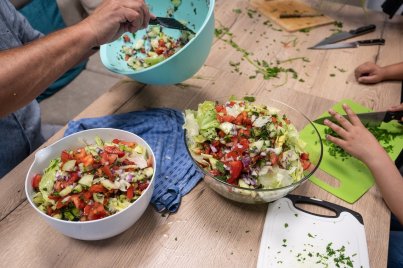 Zwei Schüsseln mit gemischtem Salat auf einem Holztisch, daneben Schneidebrett und Hände von zwei Personen.
