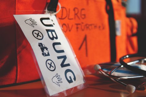 Schild mit der Aufschrift 'ÜBUNG' und Symbolen für Hände, ein Fahrzeug mit Kreuz und weitere Hände vor einem orangefarbenen Hintergrund mit unscharfem Text.