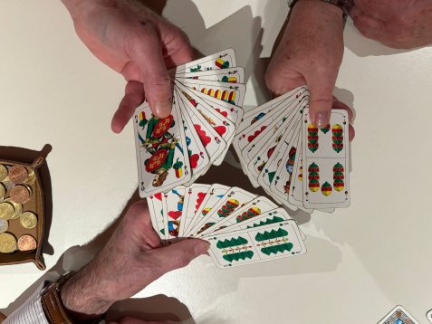 Zwei Hände halten und fächern Spielkarten mit traditionellen deutschen Symbolen über einem weißen Tisch auf, daneben eine Schale mit Münzen