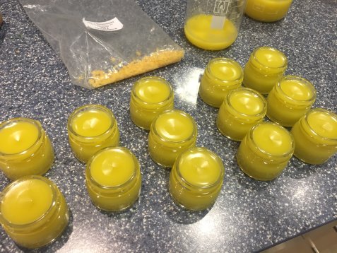 Mehrere kleine Gläser mit gelb-cremigen Salben stehen auf einer Arbeitsfläche neben einer offenen Plastiktüte mit gelben Granulatstücken