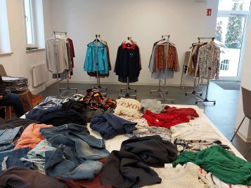 Mehrere Kleiderständer mit verschiedenen Jacken und Hemden in einem hellen Raum sowie Stapel unterschiedlicher Kleidung auf einem Tisch.