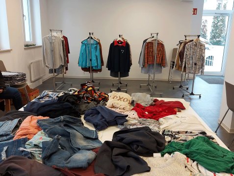 Mehrere Kleiderständer mit verschiedenen Jacken und Hemden in einem hellen Raum sowie Stapel unterschiedlicher Kleidung auf einem Tisch.