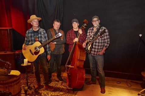 Vier Musiker mit Gitarren und Kontrabass stehen vor schwarzem Bühnenhintergrund und blicken ernst in die Kamera.