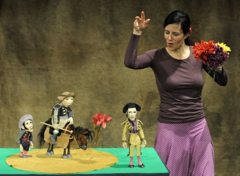 Eine Puppenspielerin steht neben einer kleinen Bühnenfläche mit Figuren aus „Ferdinand der Stier“ – darunter ein Torero, ein Junge auf einem Pferd und eine weitere Figur – und hält einen bunten Blumenstrauß in der Hand, während sie gestikulierend spielt.