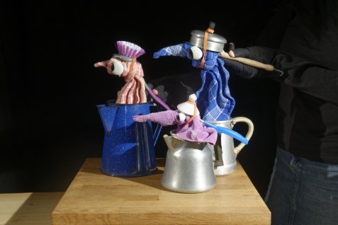 Drei bunte Stofffiguren mit großen Augen stecken in Kannen und Töpfen auf einem Holztisch, rechts ist die Hand einer spielenden Person zu sehen.