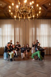 Gruppe von Menschen in schwarzen T-Shirts mit der Aufschrift 'EveryBody', einige sitzen im Rollstuhl, andere stehen oder knien in einem Raum mit Holzboden und Vorhängen