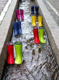 Mehrere bunte Gummistiefel stehen in einem Straßenbach.