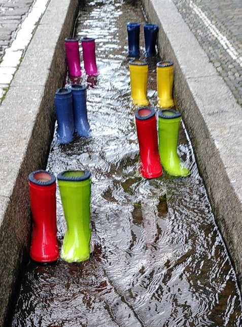 Mehrere bunte Gummistiefel stehen in einem Straßenbach.