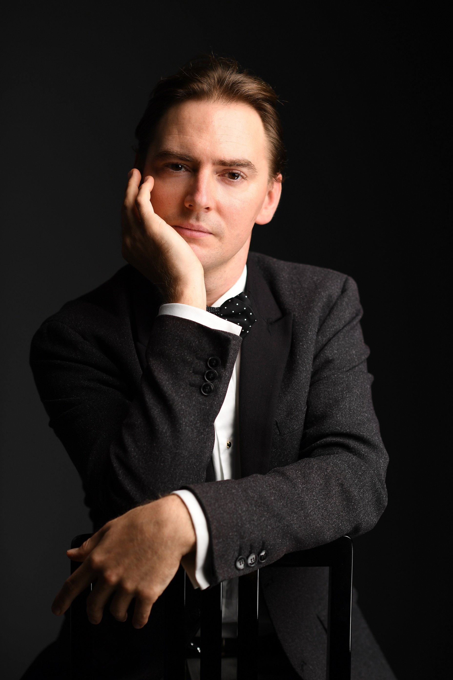 Greg Niemczuk in dunklem Anzug mit weißem Hemd und gepunkteter Fliege stützt Kopf mit Hand ab vor schwarzem Hintergrund