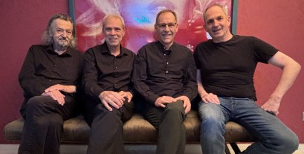 Vier Männer sitzen nebeneinander auf einer Couch, alle tragen dunkle Oberteile, einer trägt Jeans.