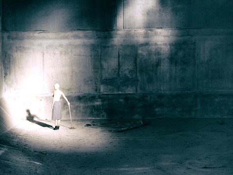 Frau mit Stock steht in einem großen, dunklen Raum im Lichtkegel an der Wand