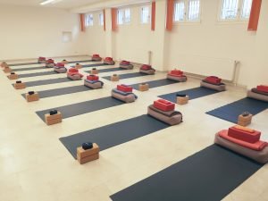 Mehrere Yogamatten mit zusammengefalteten Decken und kleinen Holzkästen in einem hellen Raum mit hohen Fenstern