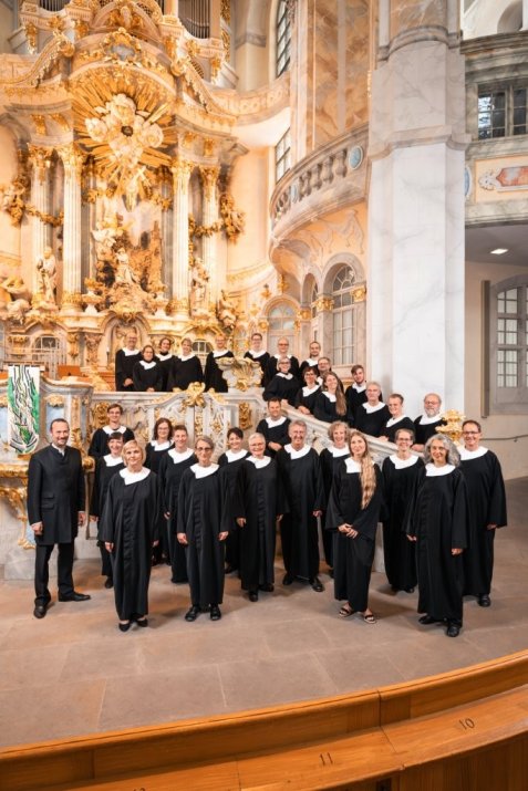 Der Kammerchor in schwarzen Roben mit weißen Kragen in einem prunkvollen barocken Innenraum mit goldverzierten Wänden und Treppe