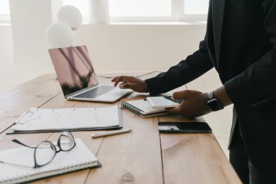 Person in dunkler Kleidung steht an einem Holztisch mit Laptop, Notizbuch, Brille und Stift