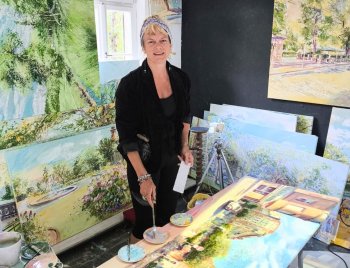 Birgit Maria Götz in schwarzer Kleidung steht in einem Raum mit mehreren Landschaftsgemälden, hält einen Pinsel und einen Zettel in der Hand