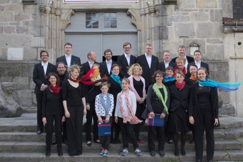 Der Chor mit Erwachsenen und Kindern vor einem historischen Gebäude, einige tragen bunte Schals und halten Ordner