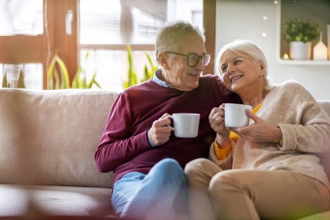 Älteres Paar sitzt eng nebeneinander auf einem Sofa und hält jeweils eine weiße Tasse in der Hand