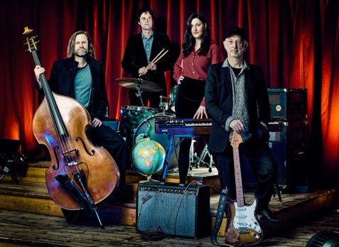 Die band mit Musikinstrumenten vor rotem Vorhang, darunter ein Kontrabass, Schlagzeug, E-Gitarre und ein Verstärker