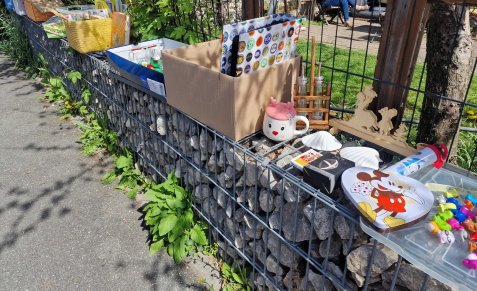 Steinmauer mit Metallgitter, darauf Kartons und verschiedene Gegenstände wie Tassen und Schachteln, im Hintergrund Garten und Gehweg