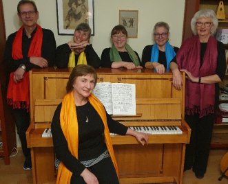 Sechs Personen mit bunten Schals stehen und sitzen um ein Klavier mit aufgeschlagenem Notenblatt