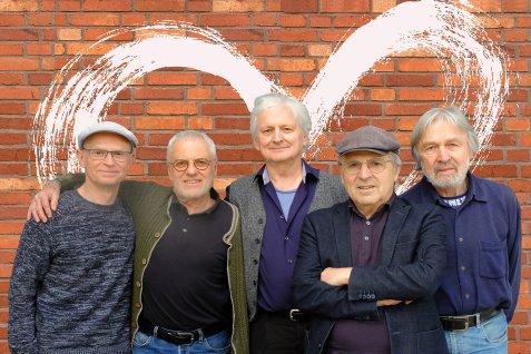 Die fünf Musiker von Young at heart stehen vor einer roten Backsteinwand, alle lächeln und haben die Arme umeinander gelegt.