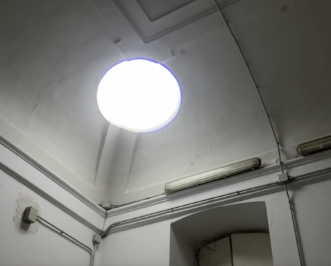 Rundes Oberlicht in einer hohen, weißen Raumdecke mit sichtbaren Kabeln und Leuchtstoffröhre an der Wand