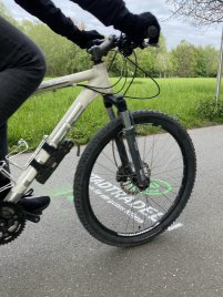Nahaufnahme eines weißen Mountainbikes mit schwarzer Bereifung und einer Person in schwarzer Kleidung, die die Lenkergriffe hält, vor einer grünen Wiese und Bäumen