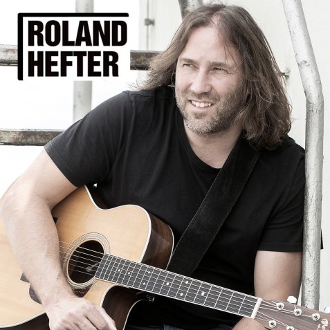 Roland Hefter mit schulterlangem Haar und schwarzem T-Shirt spielt akustische Gitarre vor einer weißen Wand mit dem Schriftzug 'ROLAND HEFTER'