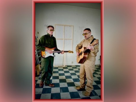 Zwei Männer in Arbeitskleidung spielen akustische und elektrische Gitarren in einem Raum mit schwarz-weiß kariertem Boden