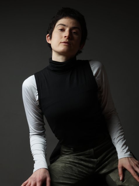 Porträt Miriam Markl mit kurzem dunklem Haar, die ein schwarzes ärmelloses Oberteil über einem weißen Langarmshirt trägt, vor dunklem Hintergrund