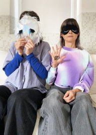 Zwei sitzende Personen tragen Schlafmasken, eine hält eine weiße Maske mit schwarzen Federn vor das Gesicht, die andere zeigt eine Hand mit einem aufgemalten Auge