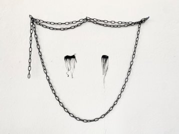 Schwarze Kette an der Wand mit zwei tropfenförmigen schwarzen Flecken darunter. Zusammen formen sie ein abstraktes Gesicht mit verlaufender Wimperntusche