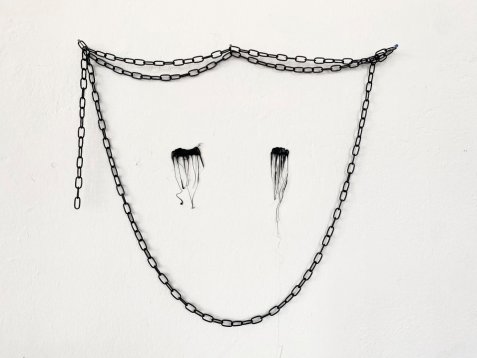 Schwarze Kette an der Wand mit zwei tropfenförmigen schwarzen Flecken darunter. Zusammen formen sie ein abstraktes Gesicht mit verlaufender Wimperntusche