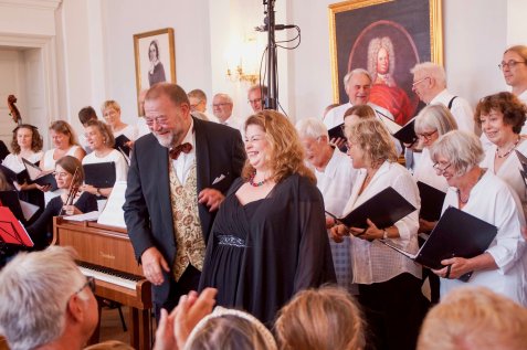 Zwei Personen in festlicher Kleidung lachen beim Schlussapplaus eines Chorkonzerts in einem eleganten Salon vor einem großen Chor.