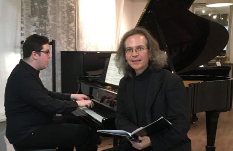 Zwei Männer in schwarzer Kleidung vor einem Flügel, einer spielt Klavier, der andere hält ein Notenheft und schaut in die Kamera