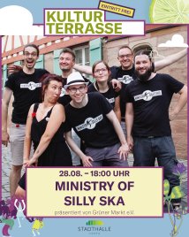 Gruppe von sieben Personen in schwarzer Kleidung mit "Ministry of Silly Ska"-Aufdrucken vor Backsteinmauer, einige lachen und haben die Arme ausgebreitet
