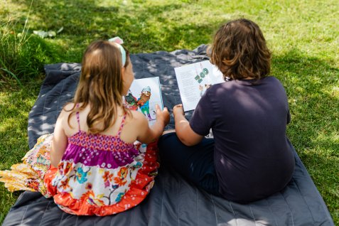 Zwei Kinder sitzen auf einer Decke im Gras und schauen gemeinsam in ein bebildertes Buch.