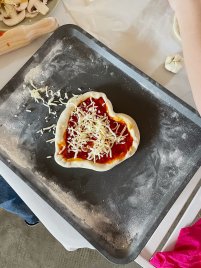Herzförmiger Pizzateig mit Tomatensauce und geriebenem Käse belegt liegt auf einem Backblech, daneben Nudelholz und Champignonscheiben.