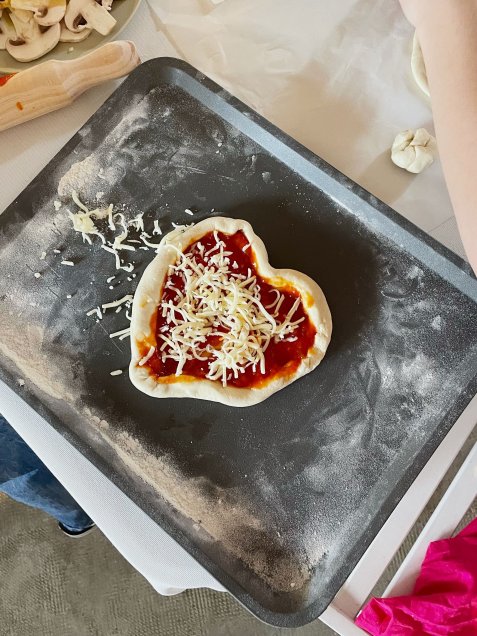 Herzförmiger Pizzateig mit Tomatensauce und geriebenem Käse belegt liegt auf einem Backblech, daneben Nudelholz und Champignonscheiben.