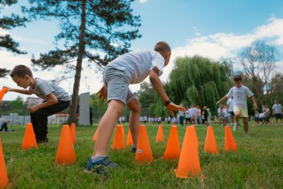 Kinder springen zwischen orangefarbenen Hütchen über eine Wiese, während im Hintergrund weitere Kinder auf einer Grünfläche versammelt sind.