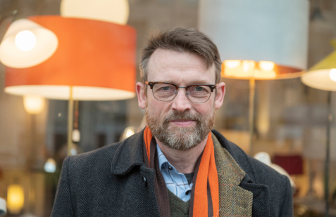 Porträt eines bärtigen Mannes mit Brille und orangefarbenem Schal vor einem Schaufenster mit beleuchteten Lampen.